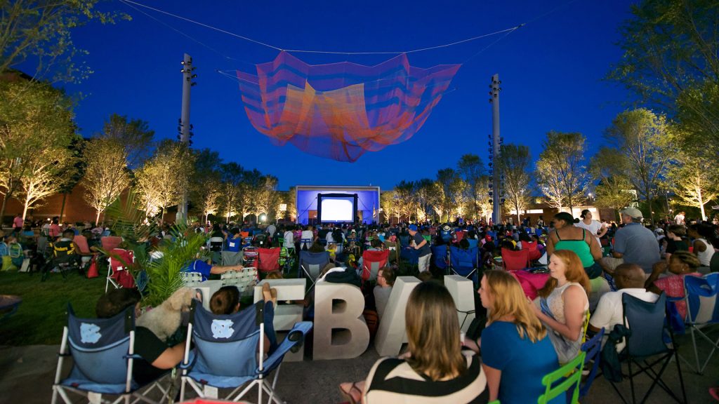LeBauer Park Open-air cinema at night with audience watching a movie beneath a colorful net sculpture, surrounded by illuminated trees.