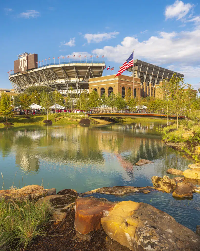 Texas A& M - Aggie Park - College Station, TX 110522 Kyle Field exterior with flag, sunny day, pond in foreground reflecting stadium and flags, surrounded by greenery.